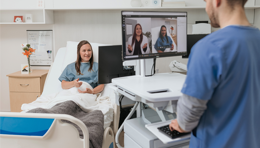 Patient sitting on bed across from a nurse on a virtual call with a doctor.