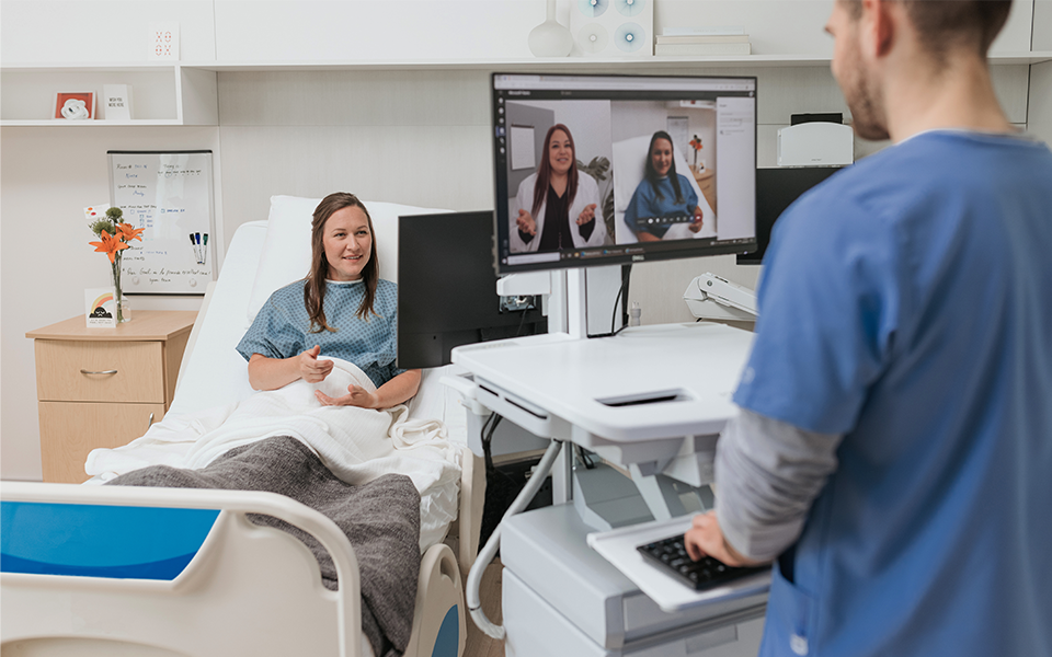 Patient sitting on bed across from a nurse on a virtual call with a doctor.